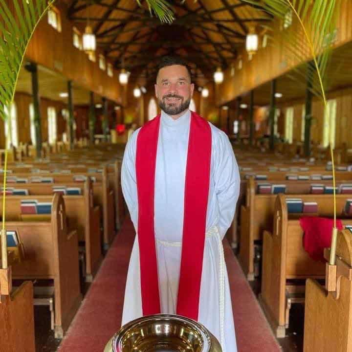 Episcopalian priest in white robe with red stoll standing and smiling in church sanctuary.